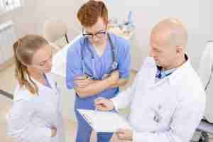 Bald serious doctor in whitecoat pointing at document while reading it aloud to young interns in clinics
